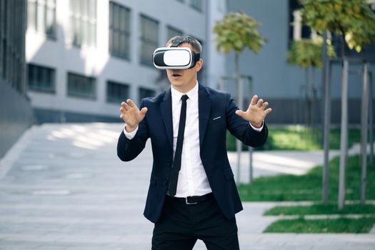 Happy businessman playing a game on imaginary screen using augmented reality goggles standing on the background of modern urban building. New technology offers new 3D dimensions.