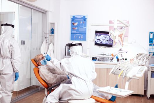 Dentist nurse taking patient temperature wearing hazmat suit using digital thermometer. Elderly woman in protective uniform during medical examination in dental clinic.