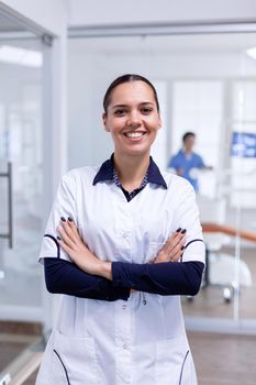 Portrait of teeth doctor in dental reception with arms crossed looking at camera wearing uniform. Happy smiling dentist in her orthodontic office.