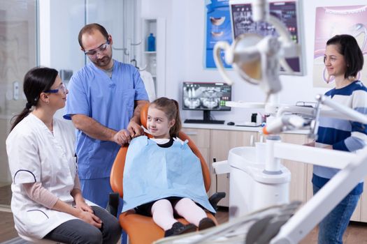Little girl wearing dental bib sitting on chair in dentist office during tooth examining .Child with her mother during teeth check up with stomatolog sitting on chair.
