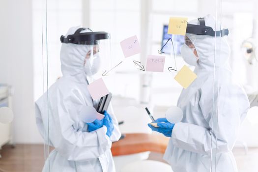 Tired dentist discussing about patient diagnosis wearing ppe suit. Medical team in stomatology office wearing coverall in dental office writing ideas on sticky notes.