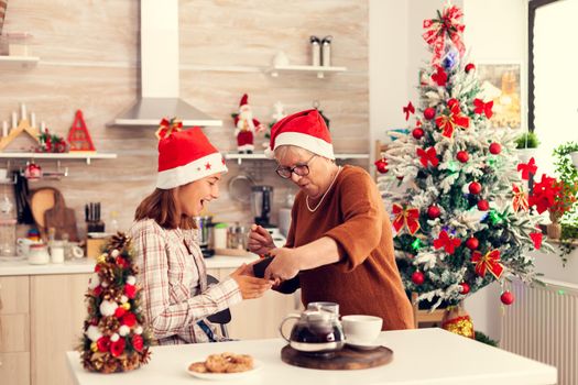 Happy grandmother giving niece gift box during christmas celebration wearing red santa hat. Senior woman wearing santa hat surprising granddaughter with winter holiday present in home kitchen with xmas tree in the background.