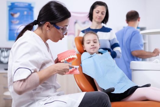 Pediatric dentist holding jaw model explaining cavity to child wearing bib. Little girl and mother listening stomatolog talking about tooth hygine in dentistiry clinic holding jaw model.