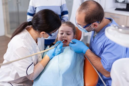 Painless treatment with modern professional dental equipment for little girl wearing bib sitting on chair. Mother with her kid in stomatology clinic for teeth examine using modern instruments.