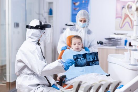 Child and mother in ppe suit listening dentist talking about dental procedure. Stomatolog in protectie suit for coroanvirus as safety precaution holding child teeth x-ray during consultation.