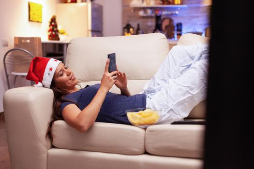 Woman lying on sofa browsing on social media using smartphone during christmastime