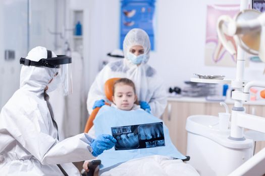 Dentist in coverall exaplaining dental procedure to kid and parent dressed in ppe suit showing radiography. Stomatolog in protectie suit for coroanvirus as safety precaution holding child teeth x-ray during consultation.