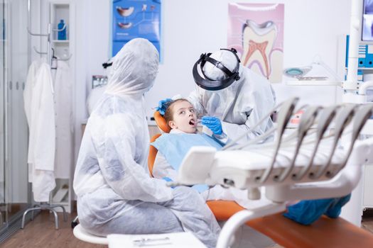 Dental specialist in the course of little girl oral hygine consultation wearing ppe suit as safety precaution because of covid19. Dentist in coronavirus suit using curved mirror during teeth examination of child.