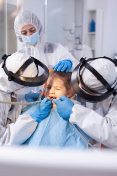 Dentist specialist treating child caries with pain wearing bib and protective suit against coronavirus. Stomatology team wearing ppe suit during covid19 doing procedure on child teeth.