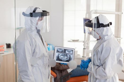 Dentist doctors with ppe suit analysing teeth x-ray using tablet in dental room, planning surgery during global pandemic while patient waiting on stomatological chair.