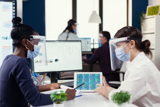 Multi ethnic employees with visor and protection mask sitting at desk