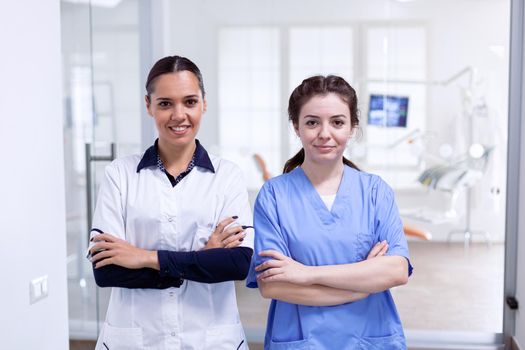 Portrait of smiling dentist and nurse smiling in stomatolog office. Portrait of stomatology team in dental reception with arms crossed looking at camera wearing uniform.