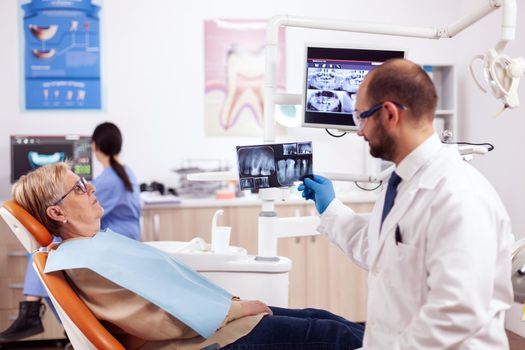 Dentist looking and examining at senior woman dental radiography for treatment. Medical teeth care taker holding patient x-ray sitting on chair during consultation.