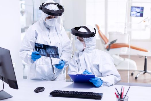 Stressed dentist and nurse examining teeth x-ray wearing ppe suit. Medical specialist wearing protective gear against coronavirus during global outbreak looking at radiography in dental office.
