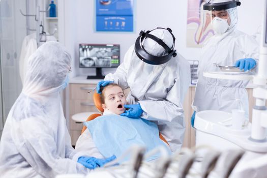 Little girl with mouth open sitting on dental chair dressed in ppe uit as safety precaution in the course of coronavirus. Dentist in coronavirus suit using curved mirror during teeth examination of child.