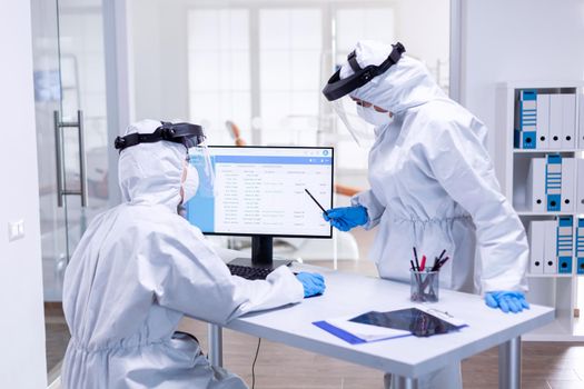 Dentist colleagues dressed in ppe suit during covid 19 using computer. Medicine team wearing protection gear against coronavirus pandemic in dental reception as safety precaution.