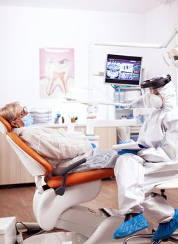 Dentist assistant wearing hazmat suit against coronavirus taking notest talking with senior patient. Elderly woman in protective uniform during medical examination in dental clinic.