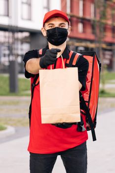 Portrait of caucasian man in red uniform, cap and medical mask from delivering service turning and smiling to camera with packet of food. Male courier with eco grocery packet