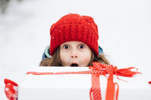 Smiling cute girl 6-7 years holding in hands birthday present in festive package on winter background. Portrait of happy girl peeking out of giftbox on Christmas day.