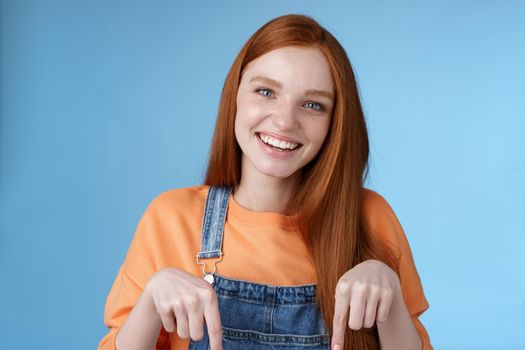 Friendly happy redhead girl smiling lively pretty grin pointing down index fingers offering good offer recommend use service standing blue background discuss interesting product, blue background