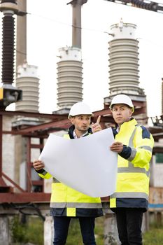 Engineers near high-voltage powerline working with a construction plan. Two engineers in special clothing discuss a drawing on paper against the background of a high-voltage power line