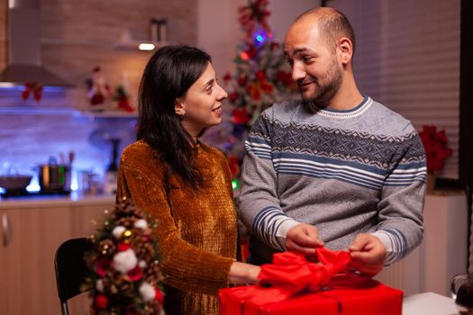 Happy couple opening xmas present gift with ribbon on it standing in x-mas decorated kitchen