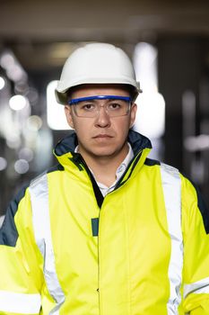 Portrait of employee serious asian man engineer worker wearing safety uniform, goggles and hardhat looking at camera on site factory warehouse background