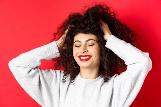 Close-up of carefree woman touching soft curly hair and smiling pleased, standing on red background. Haircare and beauty concept