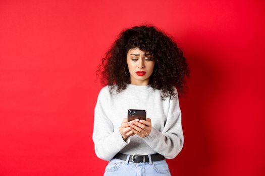 Sad and worried woman reading message on smartphone, receive bad news on social media, standing against red background
