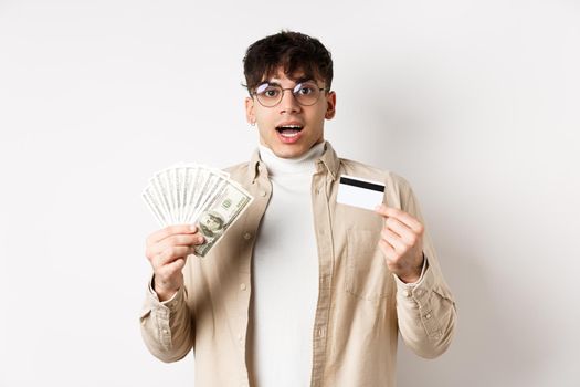 Excited young guy showing dollar bills and credit card, earn money and looking amazed, standing on white background