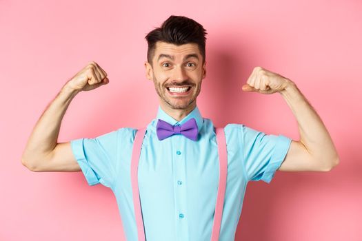 Smiling caucasian man with bow-tie and suspenders, showing muscles and feeling strong, flexing biceps to show-off, standing over pink background