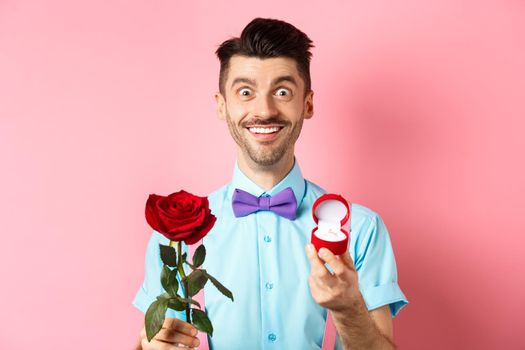 Valentines day. Smiling handsome man asking to marry him, showing engagement ring and red rose, standing romantic on pink background