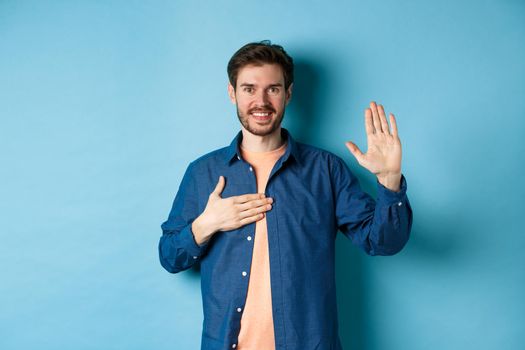 Honest young man smiling and making promise, holding hand on heart and arm raised, pledge or give oath, swearing to tell truth, standing on blue background