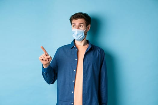 Covid-19 and healthcare concept. Surprised guy in face mask pointing and looking left at empty space, standing on blue background.