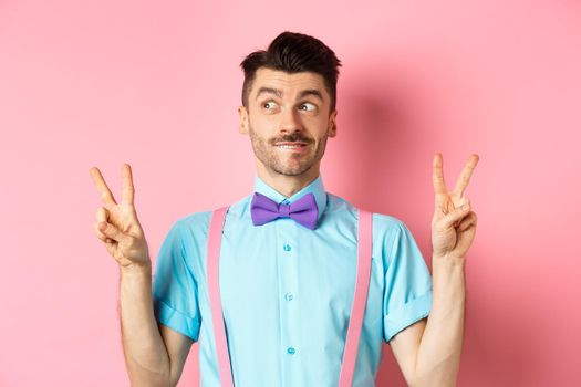 Funny young guy in bow-tie, entertain people on holiday event, showing peace signs and looking aside with excitement, standing over pink background