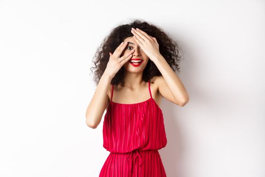 Surprise. Excited pretty girl with curly hair and red lips, peek through fingers and smiling amazed, checking out promo, standing on white background