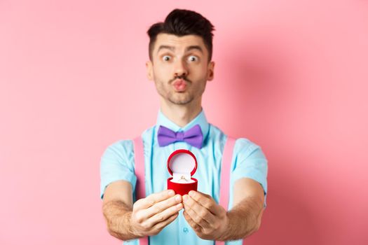 Valentines day. Funny young man pucker lips for kiss and showing engagement ring, making proposal, say marry me to lover, standing over pink background