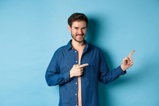 Smiling confident man showing logo, pointing fingers right at empty space and looking at camera, standing on blue background.