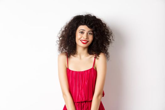 Cute modest lady in red dress and makeup, smiling and blushing, looking at camera, standing over white background