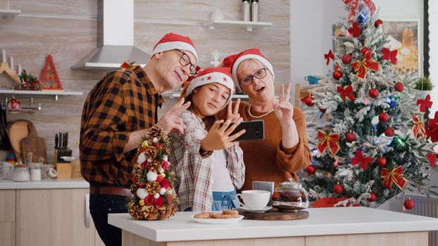 Granddaughter taking selfie using smarphone with grandparents during christmas holiday celebration in decorated kitchen. Family wearing santa hat enjoying spending time together