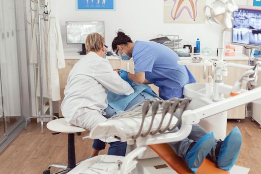 Sick man sitting on dental chair during medical examination while senior dentist woman doing oral surgery in dentistry office. Hospital team examining patient toothache preparing tooth treatment