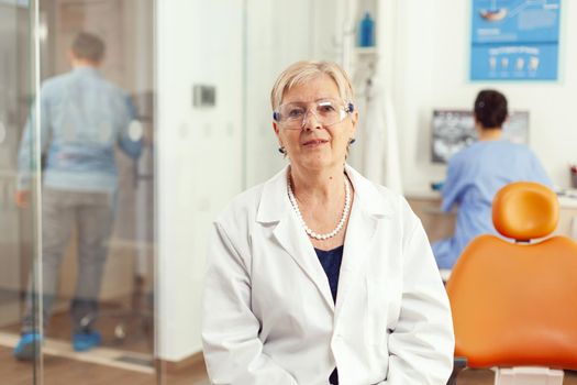 Portrait of specialist senior dentist woman in stomatological office while medical assistant discussing with patient in background. Stomatologist doctor looking on webcam