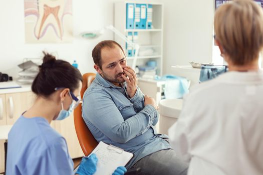 Man patient with tooth pain explaining dental problem to nurse during orthodontic appointment. Assistant writing notes on clipboard while doctor dentist checking stomatology radiography