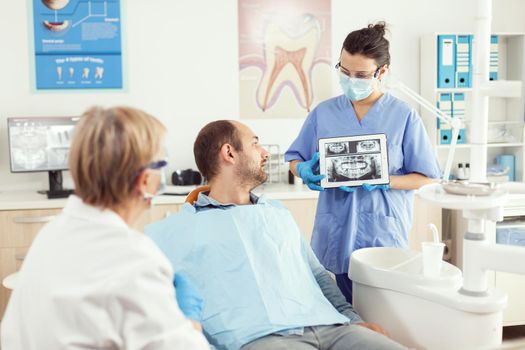 Dentist woman in dental office discussing with sick man while nurse showing tooth x-ray image on tablet. Man patient waiting toothache treatment sitting on dental chair during medical examination