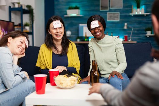 Mixed race friends playing Guess who game with sticky papers attached on forehead. Group of multiracial people having fun, laughing together while sitting on sofa in living room late at night.