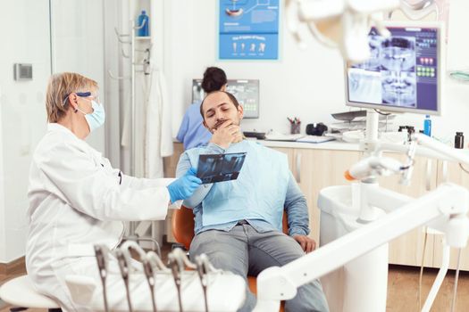 Senior woman dentist examining tooth radiography with sick man discussing toothache during stomatology appointment. Patient sitting on dental chair in hospital dentistry office