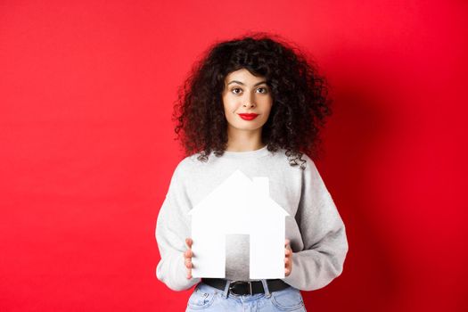 Real estate. Young caucasian woman in casual clothes showing paper house cutout, buying property or renting apartment, standing on red background
