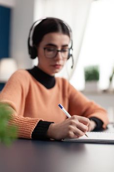 Young college student writing communication information on notebook for online school exam using e-learning platform. Student with headphone doing homework while sitting at desk table in living room
