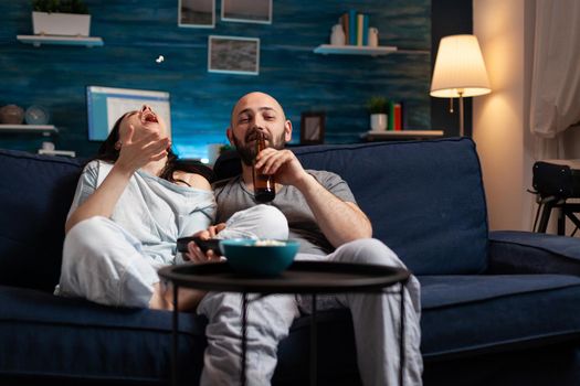 Relaxed couple in pajamas relaxing on sofa eating popcorn watching comedy movie