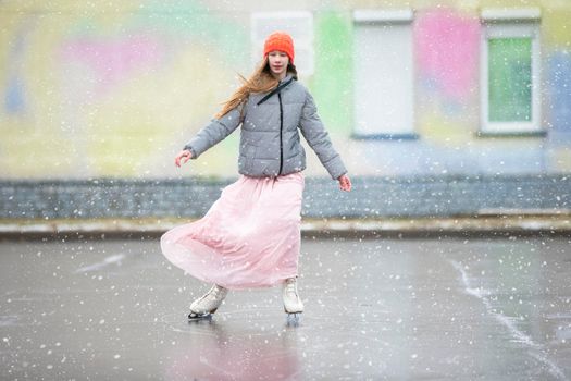 Girl teenager skates on ice in the city.Young girl in a long dress and a warm jacket on the ice.